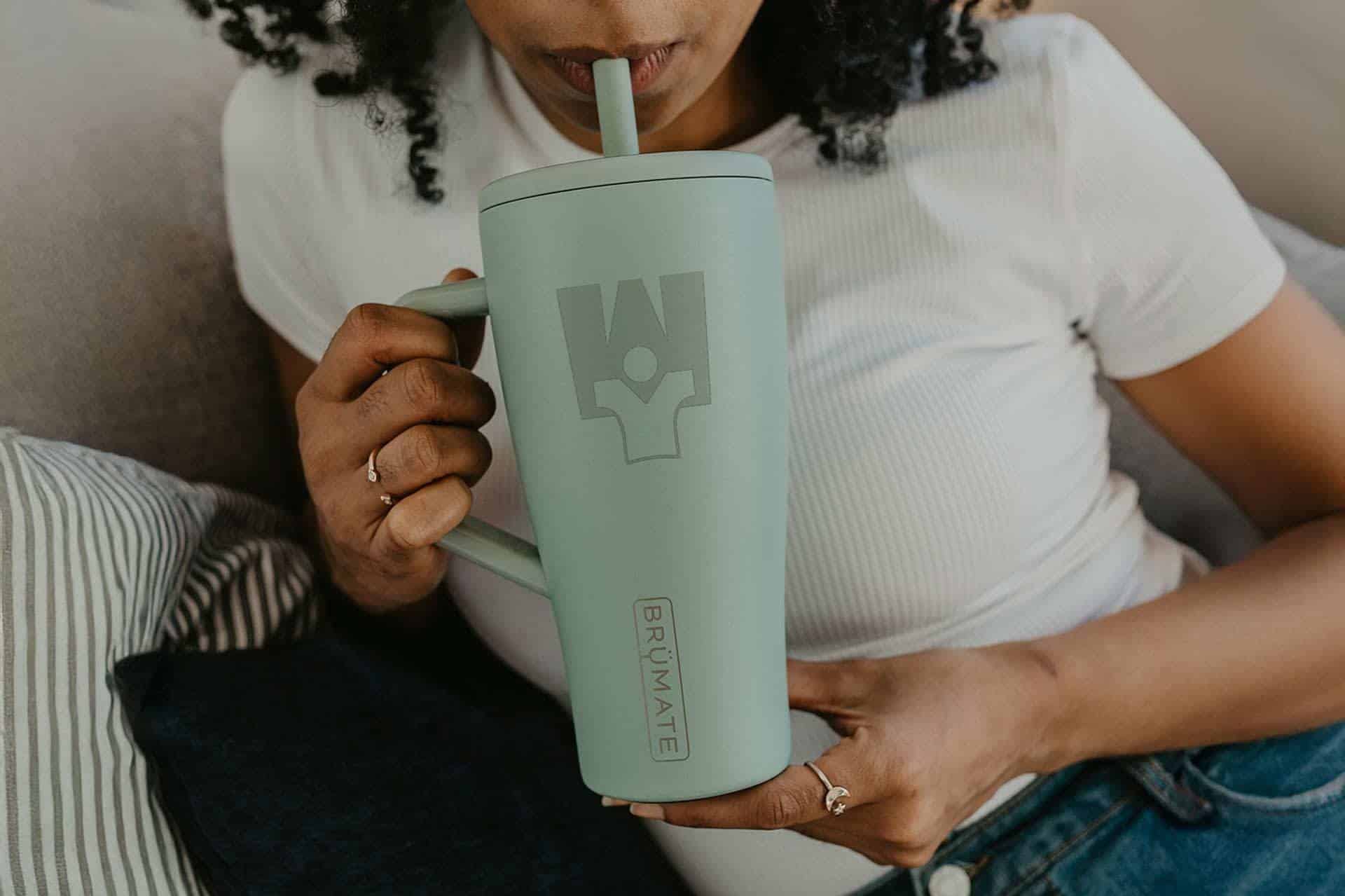 Person drinking from a large green mug with Brümate logo, sitting on couch.