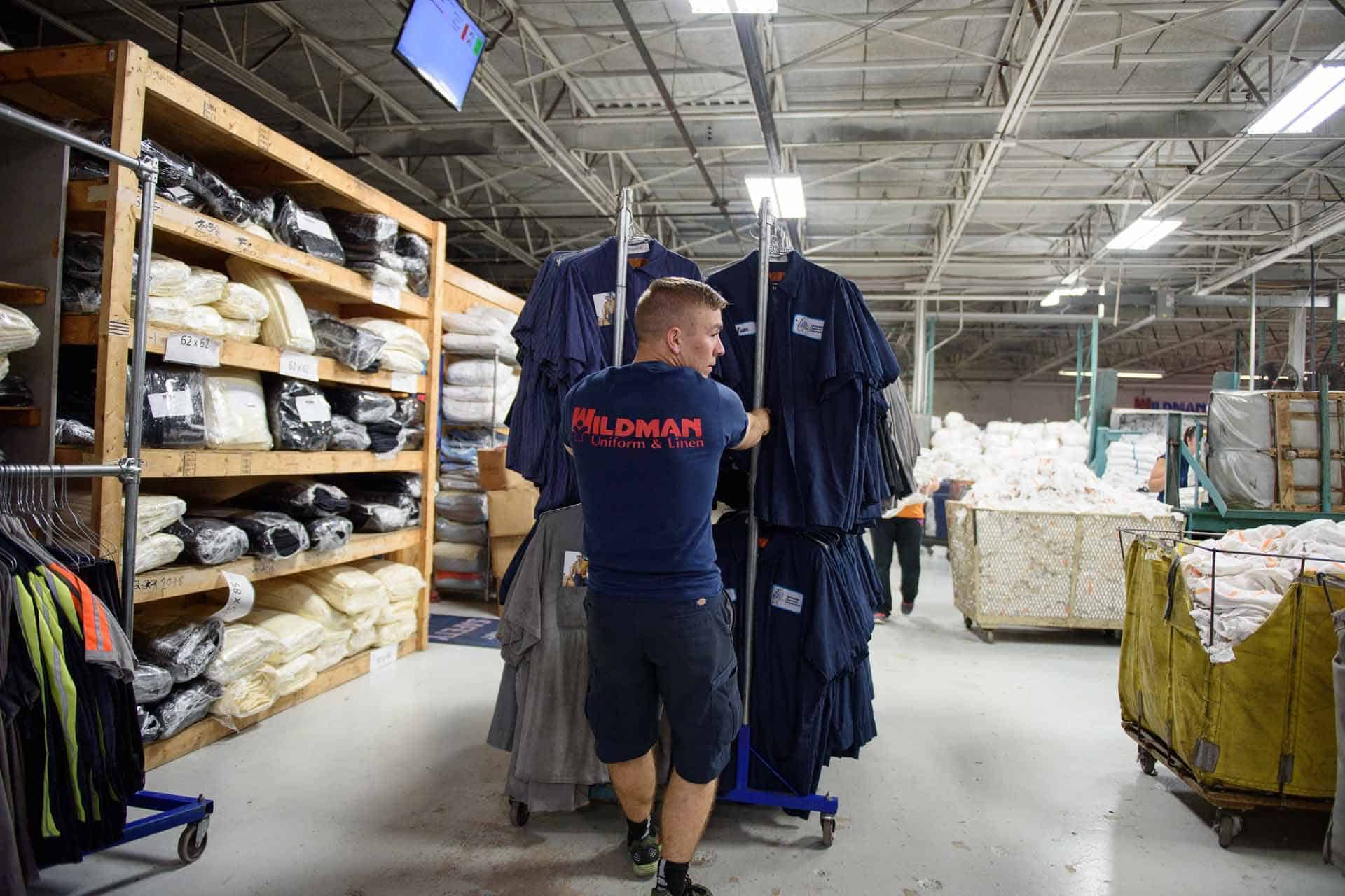 Worker organizing uniforms on racks in an industrial warehouse setting.