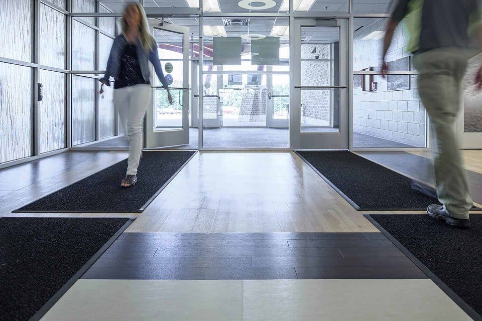 Two people walking through a modern building entrance with floor mats.