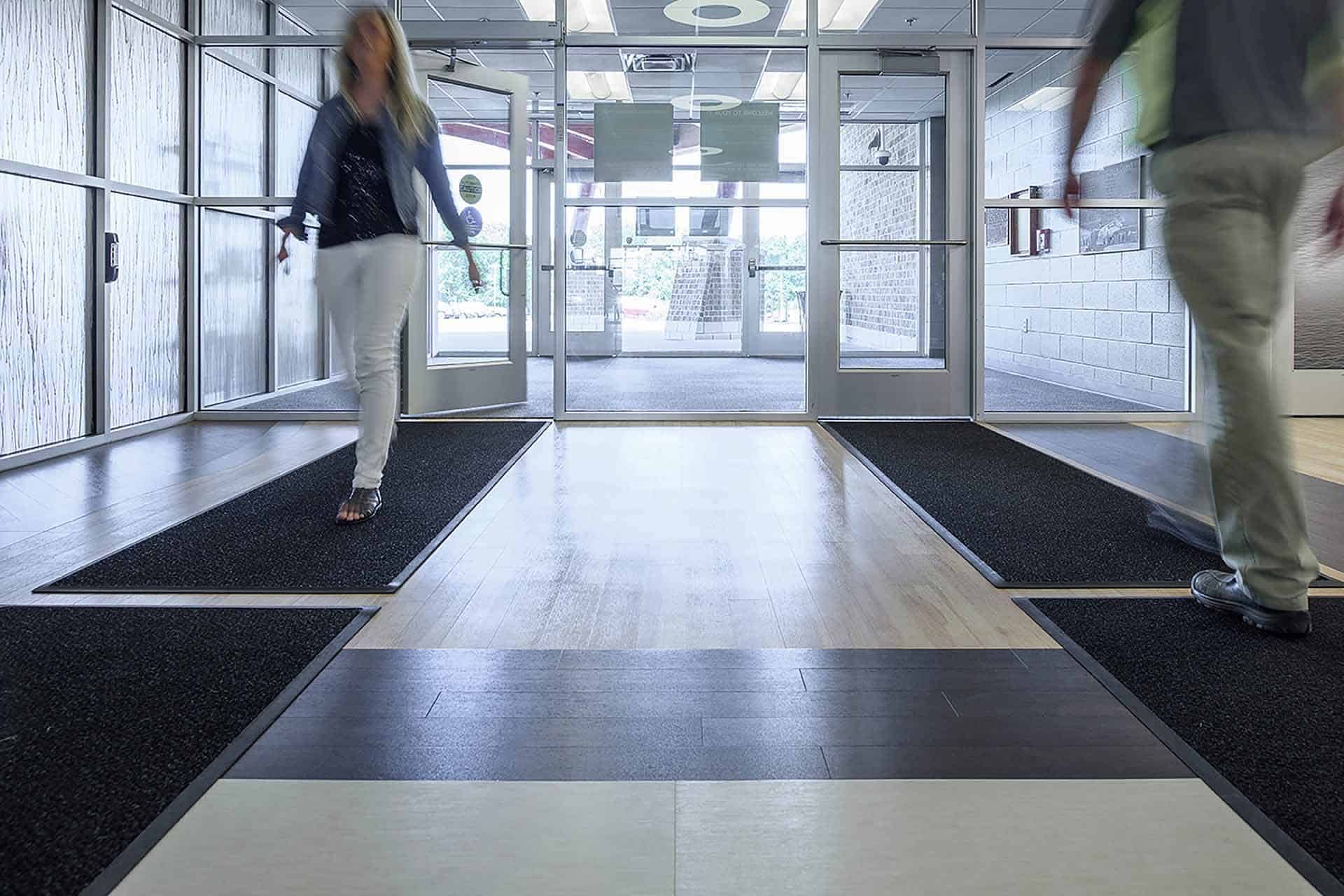 Two people walking through a modern building entrance with floor mats.