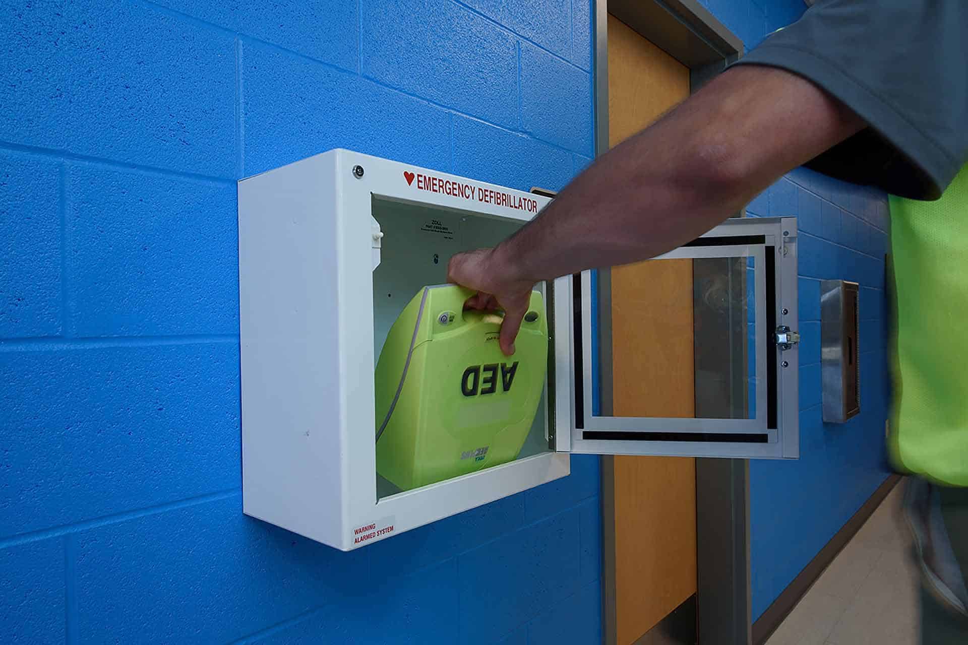 Person retrieving AED from a wall-mounted emergency defibrillator cabinet.