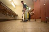 A man cleans a tiled restroom floor with a vacuum-like tool.