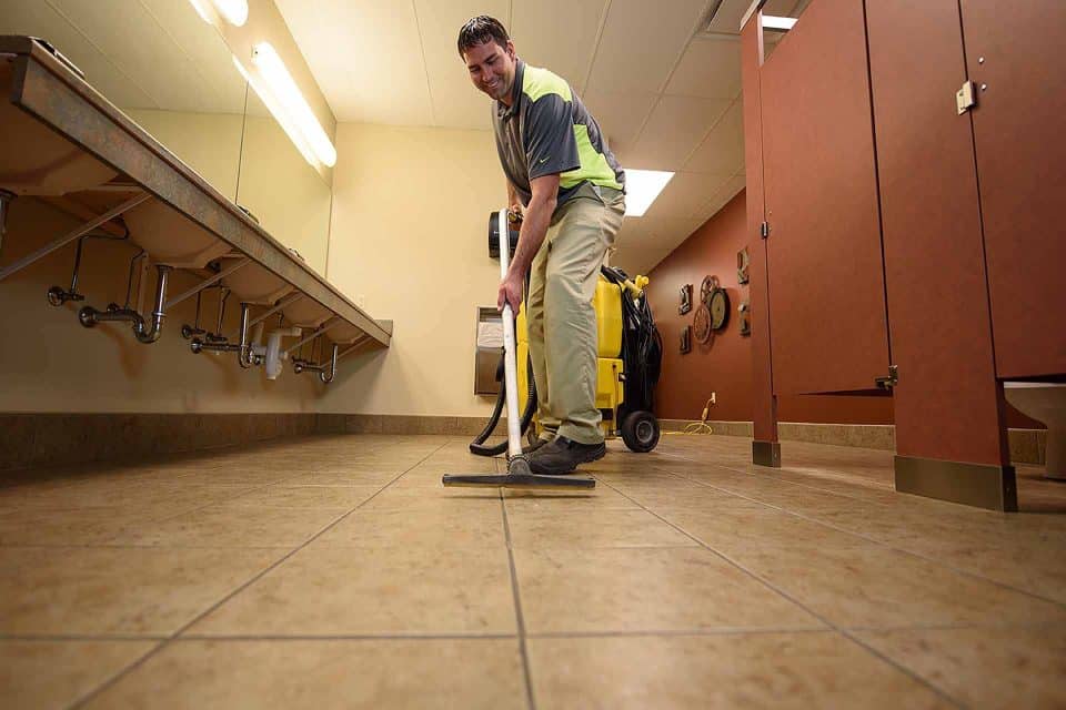 A man cleans a tiled restroom floor with a vacuum-like tool.