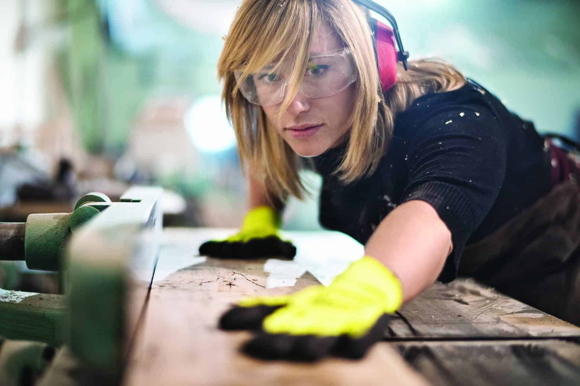 Woman wearing safety gear sanding wood in a workshop.