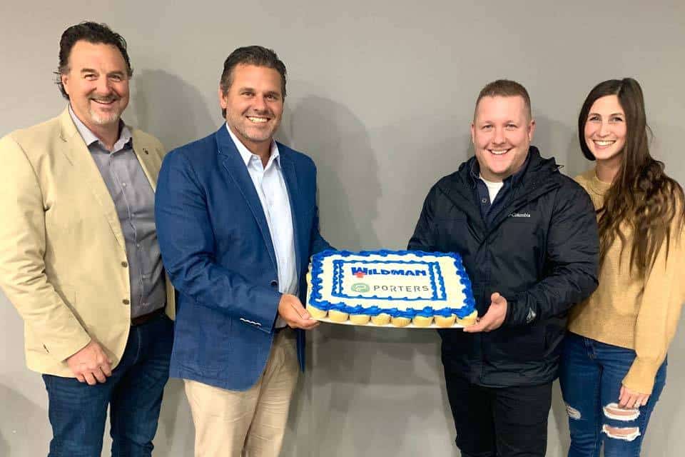 Four people standing together, two holding a rectangular cake with WILDMAN text.