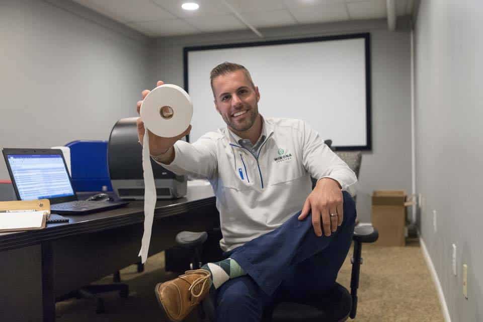 Man sitting at a desk holding up a roll of toilet paper in an office.