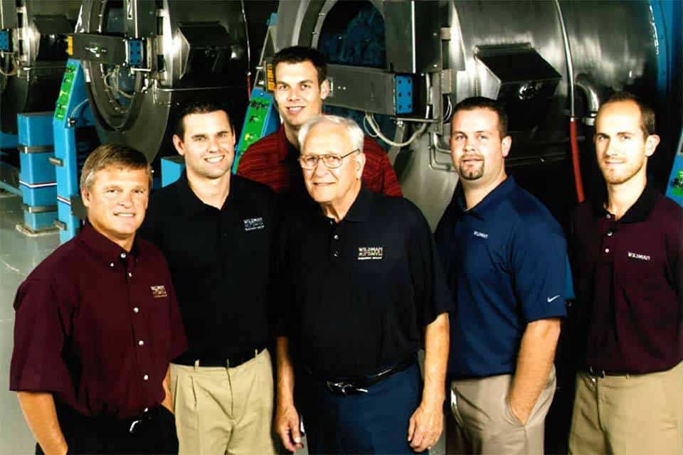 Six men stand in front of large industrial machines, posing for a group photo.