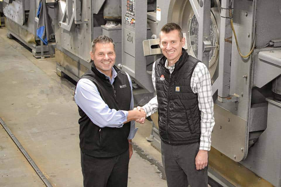 Two men in vests shake hands in an industrial facility.