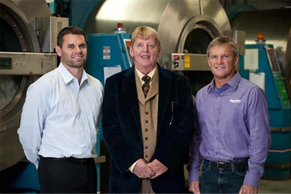 Three men stand in front of large industrial washing machines in a laundry facility.