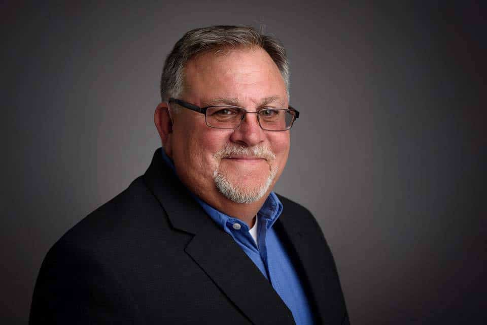 Man in glasses wearing a black blazer and blue shirt, posing against a gray background.