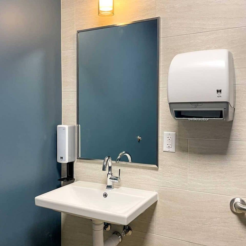 A restroom sink with a mirror, soap dispenser, paper towel dispenser, and light.