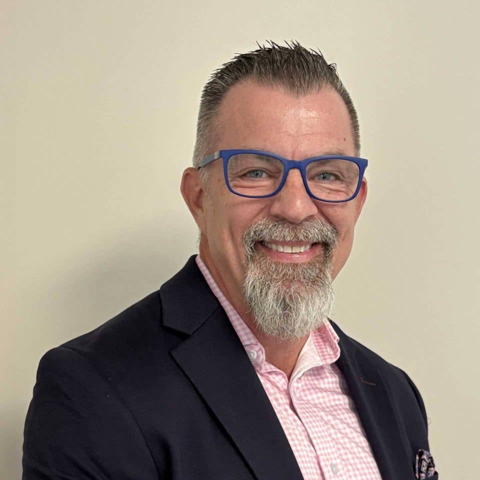 Man with short hair, blue glasses, and beard in a suit, smiling at camera.