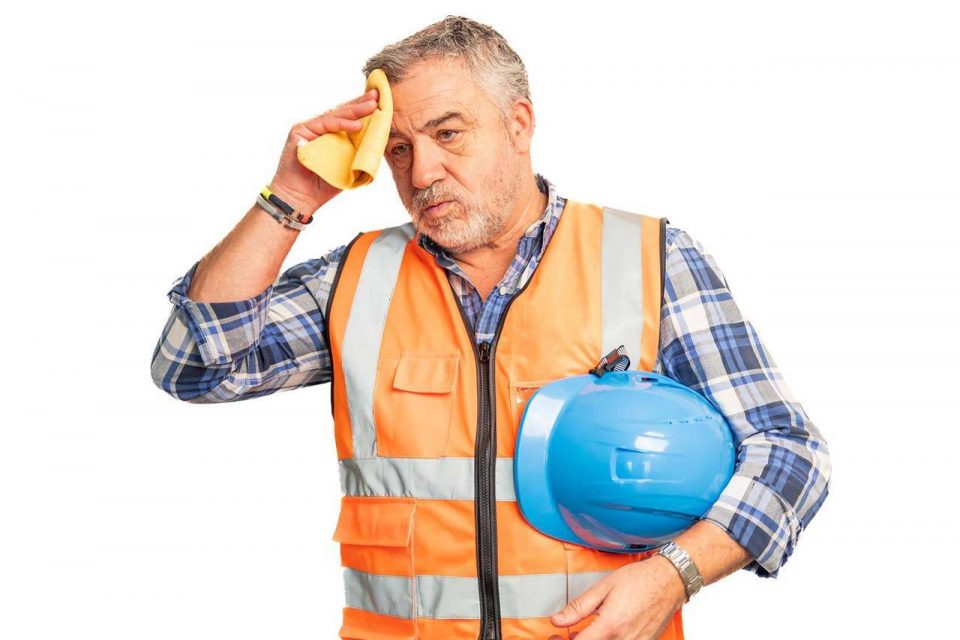 Construction worker in safety vest wipes his forehead while holding a blue helmet.