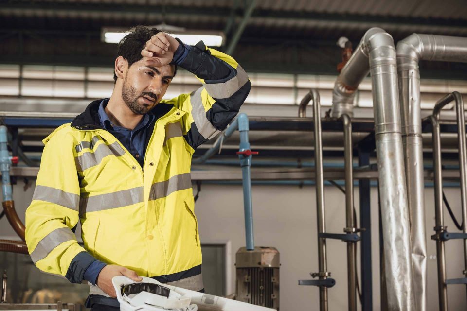 Man in a yellow safety jacket wipes sweat in an industrial facility.
