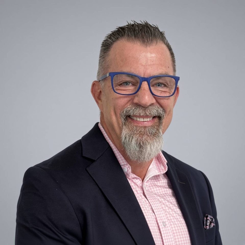A smiling man with a goatee and blue glasses wearing a navy blazer.