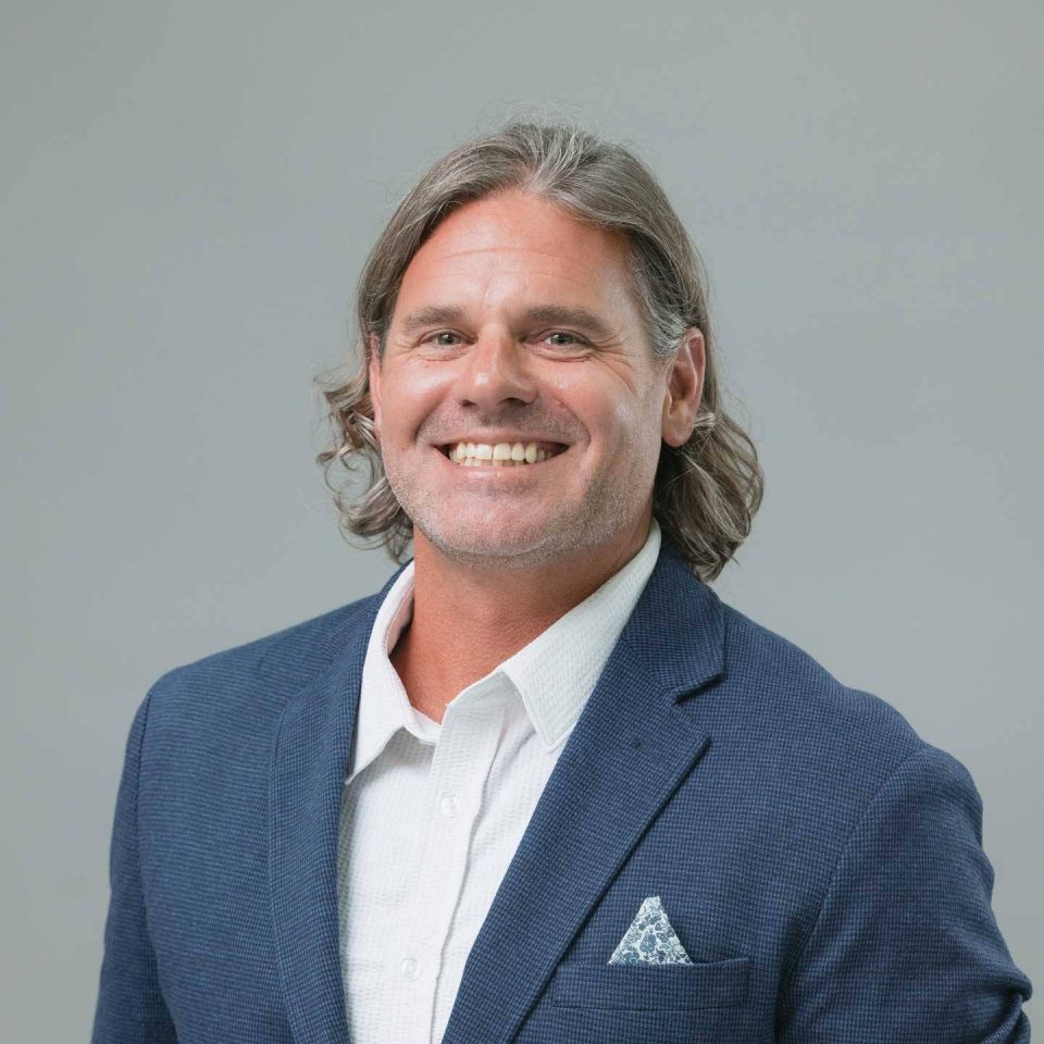 A smiling man with shoulder-length hair wearing a blue blazer and white shirt.